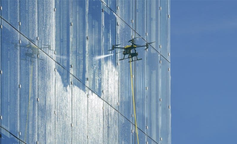 Drone soft washing the glazed elevation of a high-rise tower, tethered supply line visible